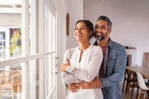 Smiling adult couple hugging each other and standing near window while looking outside.
